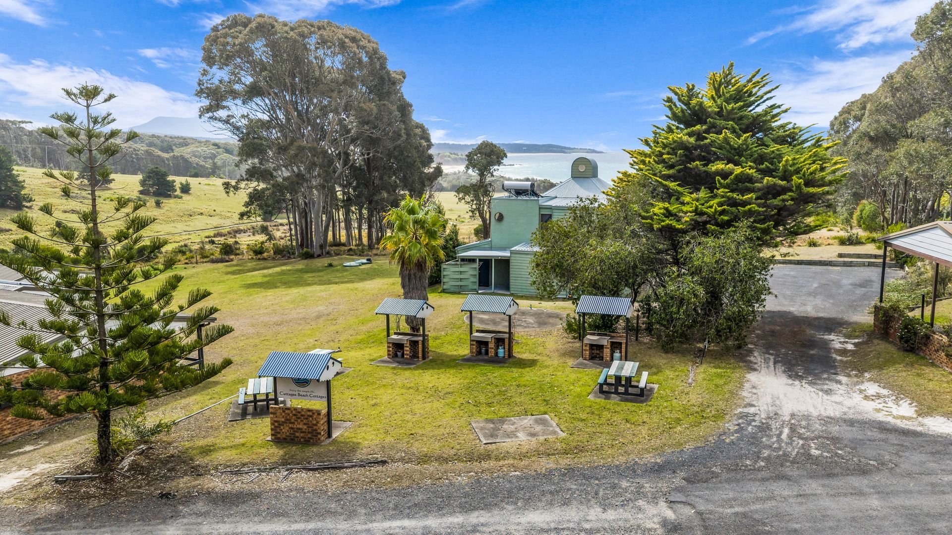 A cottage with a green roof, surrounded by a grassy area with picnic tables and a view of the sea.