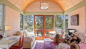 A cozy room with a wooden ceiling, a large window, and a view of trees. There is a sofa, a chair, and a small table with a lamp.