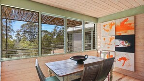 A dining area with a wooden table and chairs, large windows with a view of trees, and a painting on the wall.