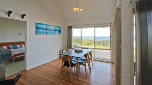 Une pièce avec un plancher en bois, un lit recouvert d’une courtepointe à rayures, une table à manger avec des chaises, un lustre et une grande fenêtre donnant sur l’océan.