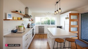 A modern kitchen with white cabinetry, a large island, bar stools, and a view of greenery outside. - Blue Pool Hideaway - Scenic Drive 7 (Bermagui)