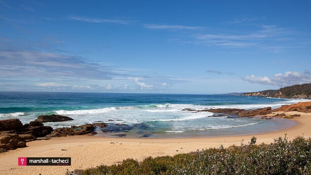 A beach with clear skies, rocky shoreline, and a sign for 'marshall + tacheci real estate'. - Blue Pool Hideaway - Scenic Drive 7 (Bermagui)