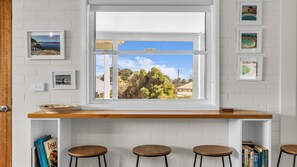 A room with a white brick wall, a window with a view of trees and a clear sky, a wooden shelf with books, and three round stools. - Blue Pool Hideaway - Scenic Drive 7 (Bermagui)