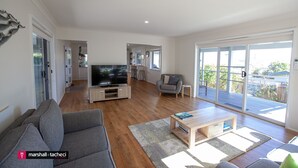 A modern living room with a grey sofa, a wooden coffee table, and a flat-screen TV. There is a sliding glass door leading to a balcony. - Blue Pool Hideaway - Scenic Drive 7 (Bermagui)
