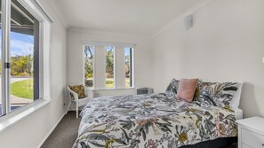 Een slaapkamer met een groot raam, een bed met bloemenbedrukking, een witte stoel en een radiator.