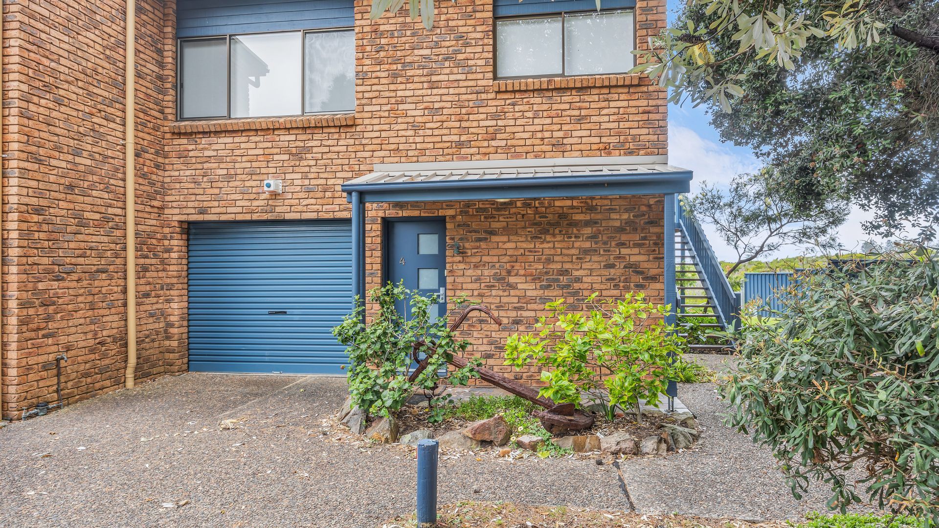A two-story brick building with a blue garage door, a covered porch, and a small garden area with plants and rocks.