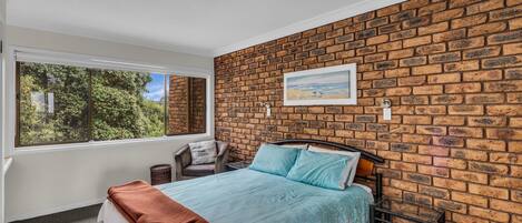 Een slaapkamer met een bakstenen muur, een bed met blauw beddengoed, een raam met uitzicht op bomen en een schilderij aan de muur.