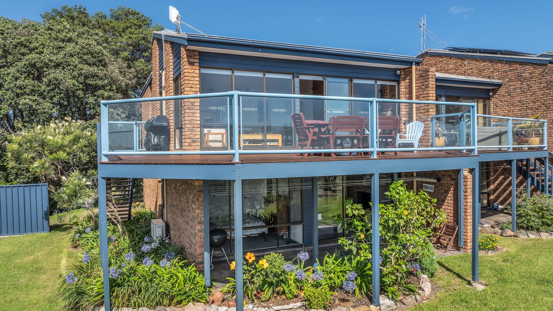 A two-story house with a glass balcony, outdoor seating, and a garden.