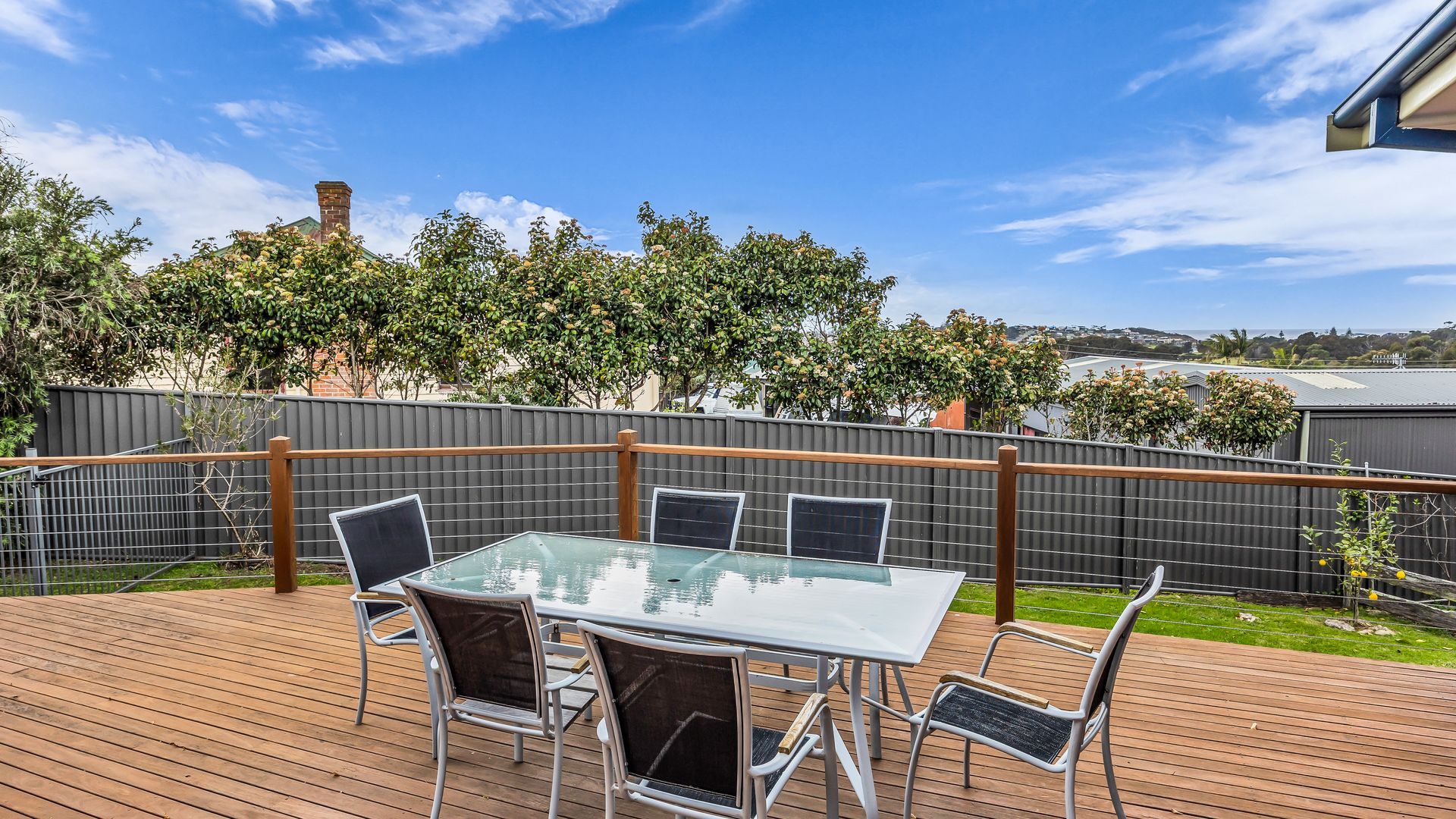 A deck with a glass-top table and chairs, surrounded by a wooden fence, with trees and a clear sky in the background.