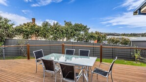 A deck with a glass-top table and chairs, surrounded by a wooden fence, with trees and a clear sky in the background.