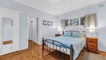 A bedroom with a wooden floor, a metal bed frame, a bedside table with a lamp, and a window with curtains.