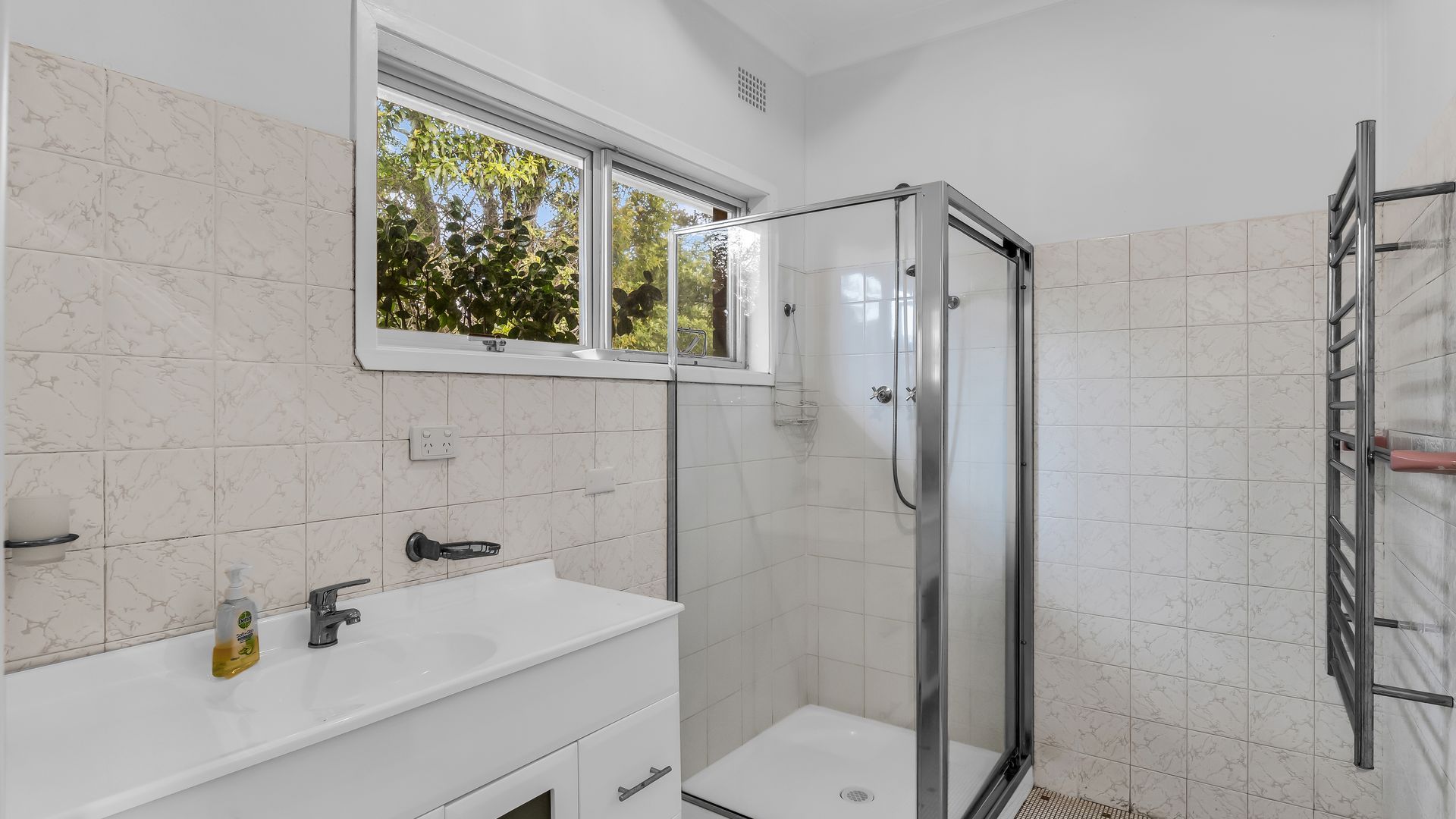 Une salle de bain moderne avec une douche à parois de verre, un lavabo blanc et une fenêtre donnant sur la verdure.