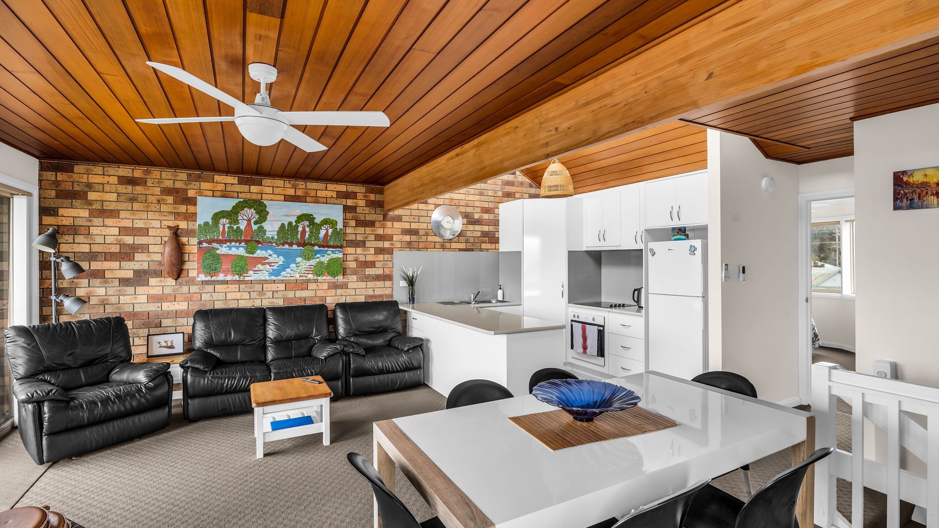 A modern living room with a brick wall, leather sofas, a dining table, and a kitchen area with white cabinets.