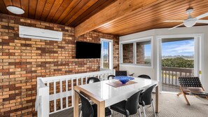 Een overdekte patio met een bakstenen muur, een flatscreen televisie, een eettafel met stoelen en een plafondventilator.