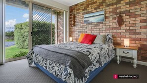 A bedroom with a large bed, a nightstand, a lamp, and a view of a garden through a sliding glass door. - Marina Townhouse 1 - Unit 1 Wapengo St 6 (BERMAGUI)