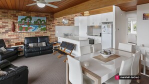 A modern living room with a black leather sofa, a dining table with white chairs, and a kitchen area with white cabinets and appliances. - Marina Townhouse 1 - Unit 1 Wapengo St 6 (BERMAGUI)
