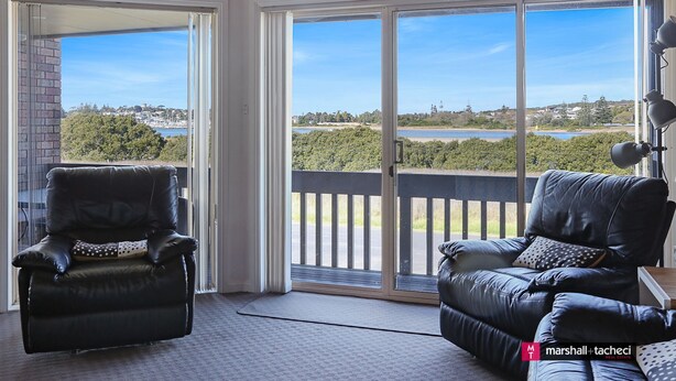 A room with a sliding glass door leading to a balcony with a view of a body of water and greenery. There are two black leather chairs with cushions. - Marina Townhouse 1 - Unit 1 Wapengo St 6 (BERMAGUI)