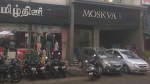 A building with a sign for 'MOSKVA HOTEL' and another sign for 'SOM'. There are parked cars and motorcycles in front of the building. - Moskva Hotel (Madurai)