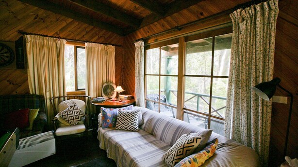 Una habitación de cabaña acogedora con techo de madera, un sofá con cojines estampados, una silla, una mesa con libros y una lámpara, y ventanas grandes con cortinas.