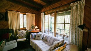 A cozy cabin room with a wooden ceiling, a sofa with patterned cushions, a chair, a table with books and a lamp, and large windows with curtains. - Nutley Cottage - Nutleys Creek Rd 74 (Bermagui)