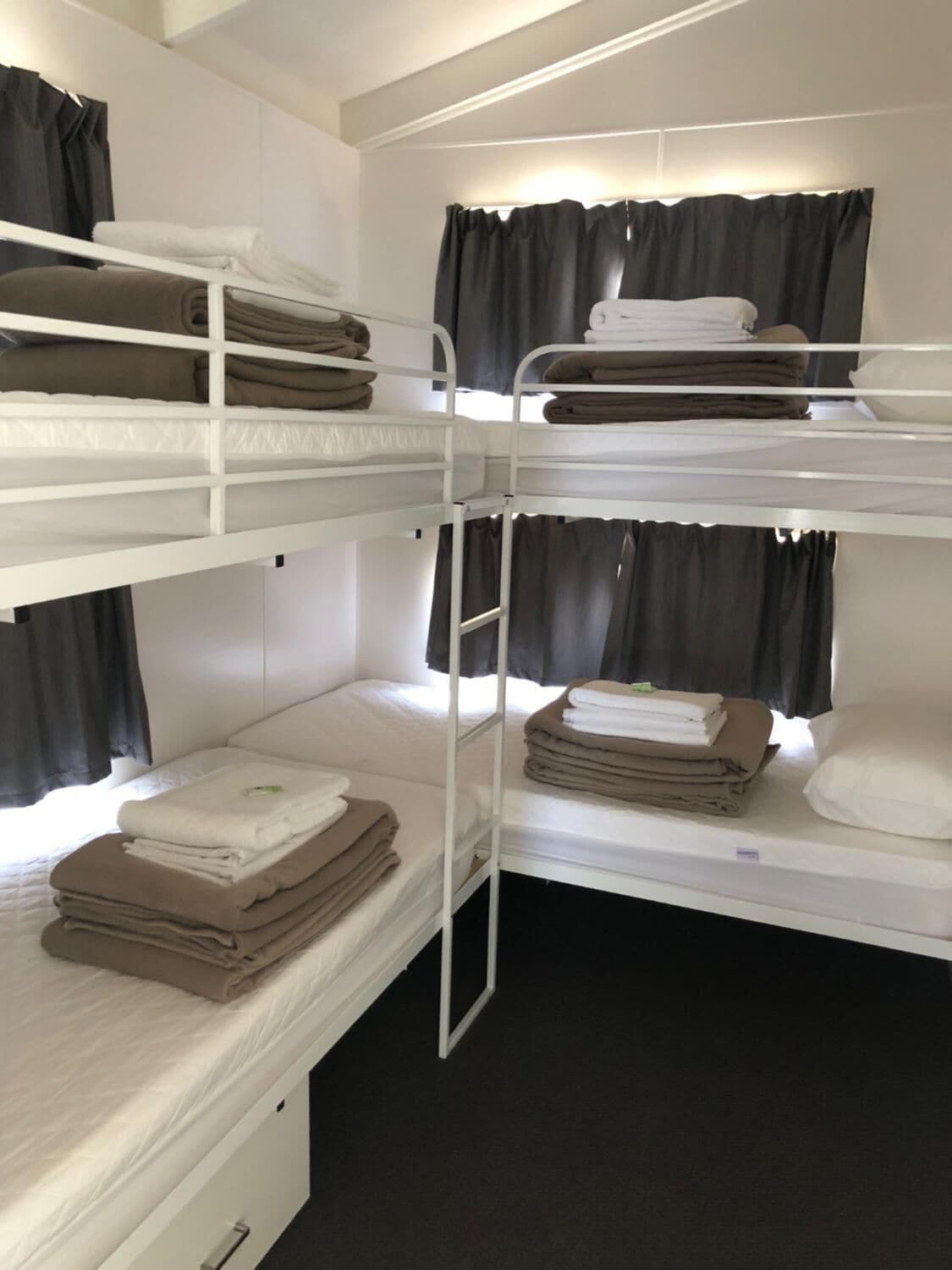 Een slaapkamer met stapelbedden, witte metalen hekken, zwarte gordijnen en netjes opgemaakte handdoeken op elk bed.