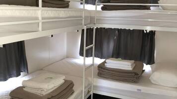Een slaapkamer met stapelbedden, witte metalen hekken, zwarte gordijnen en netjes opgemaakte handdoeken op elk bed.