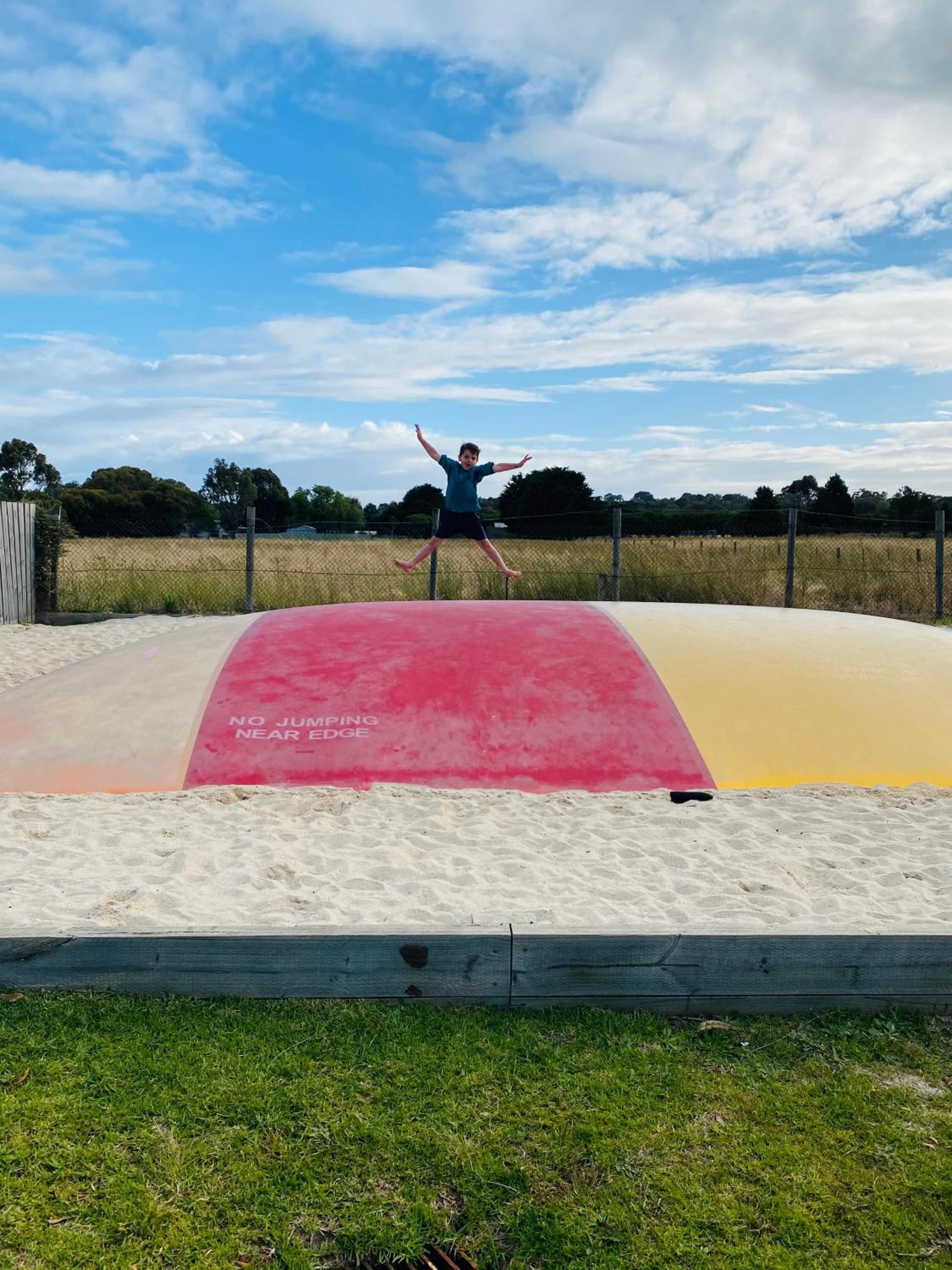 Iemand springt op een trampoline nabij een strand, met een waarschuwingsbord waarop staat: 'NO JUMPING NEAR EDGE'.