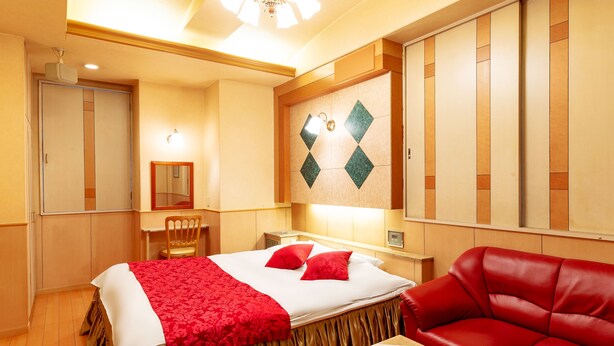 Une chambre d’hôtel avec un lit, un canapé en cuir rouge, un bureau avec une chaise, un miroir et un mur orné d’un motif géométrique.