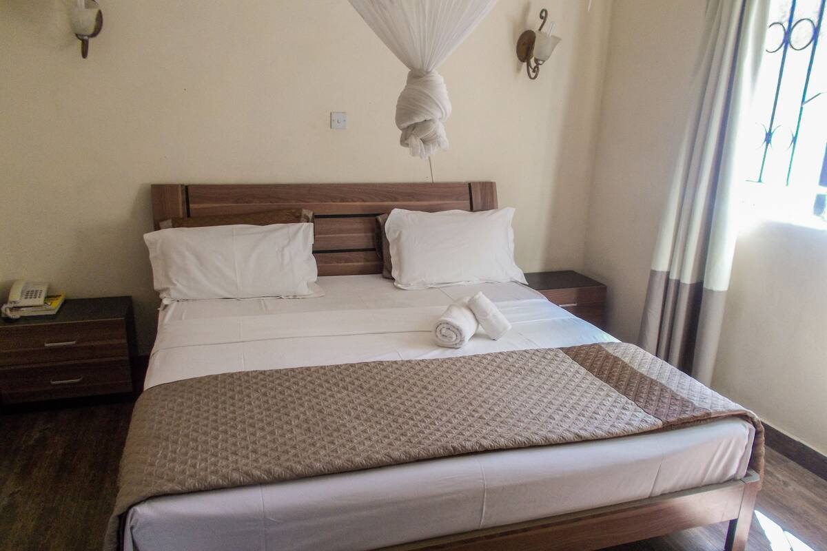 Un lit bien fait, avec une tête de lit en bois, du linge de lit blanc et une serviette enroulée posée sur le lit. On aperçoit une table de chevet sur laquelle se trouvent un téléphone et une lampe.