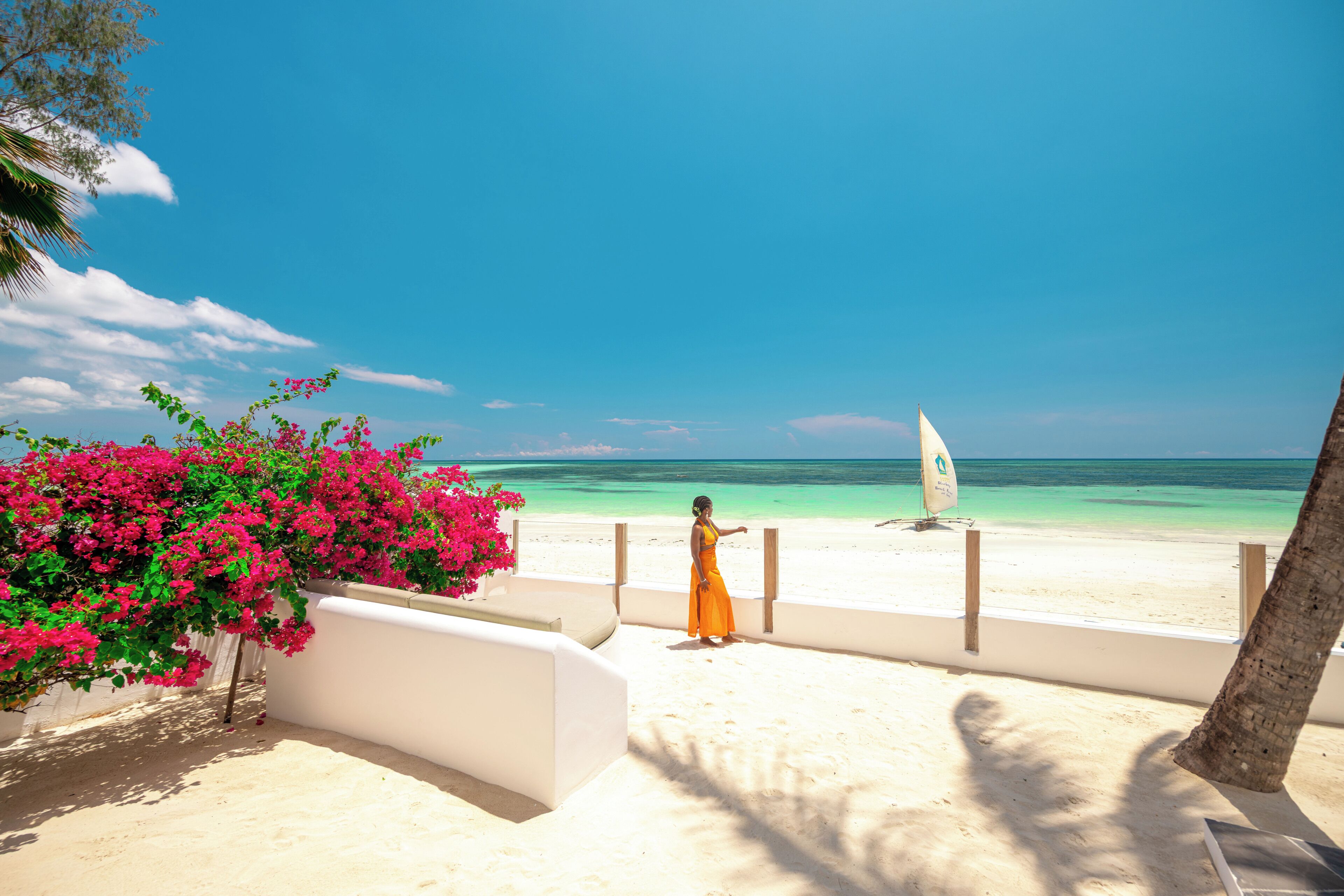 A beach view with a person in an orange dress, a white sailboat, and pink flowers.