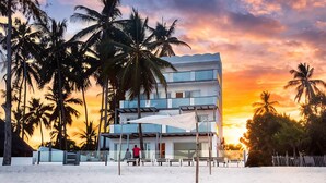 Uma praia com desenho de areia em formato de coração, palmeiras e um edifício moderno com grandes janelas.