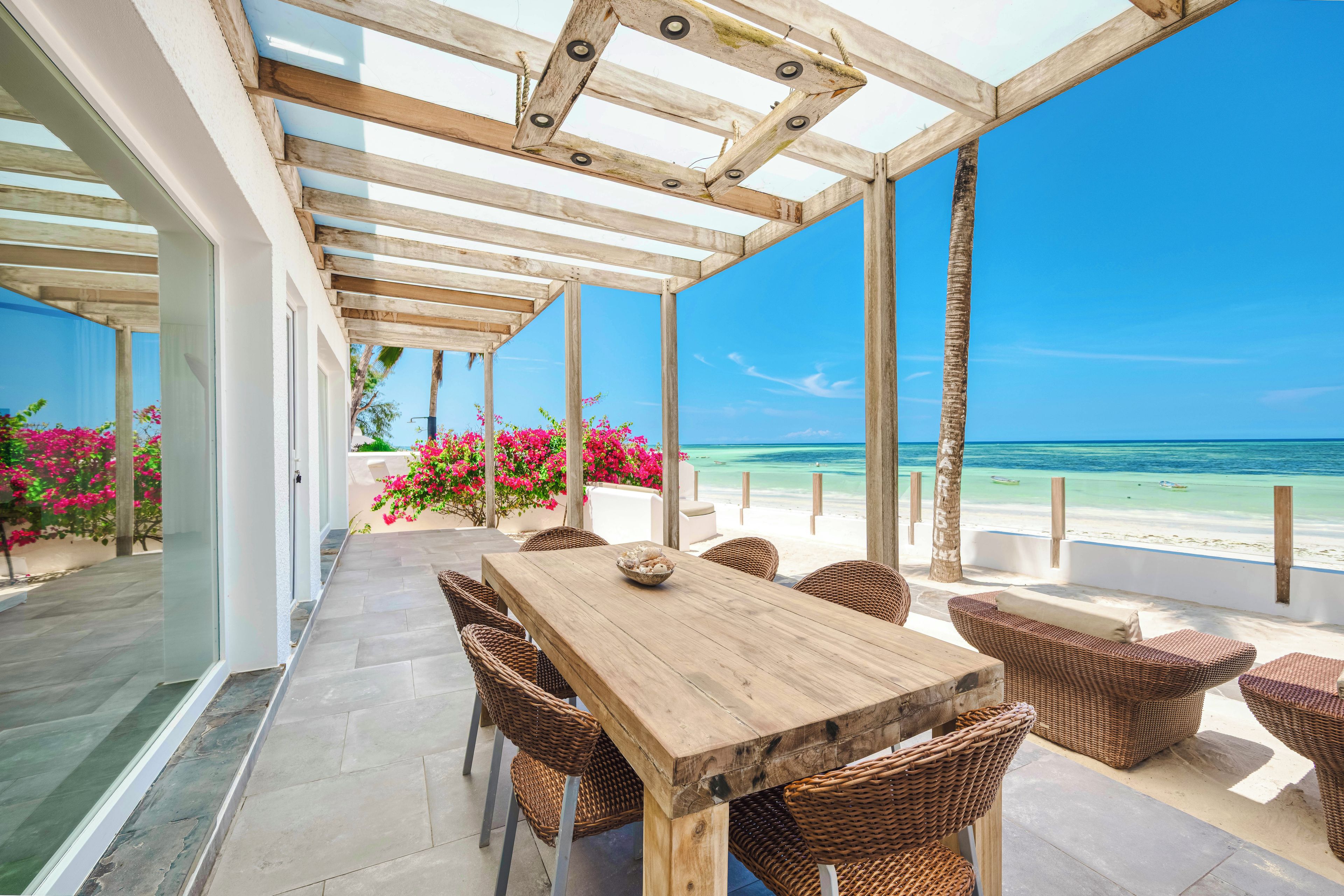 An outdoor patio with a wooden table and wicker chairs, overlooking a beach with clear blue water and pink flowers.