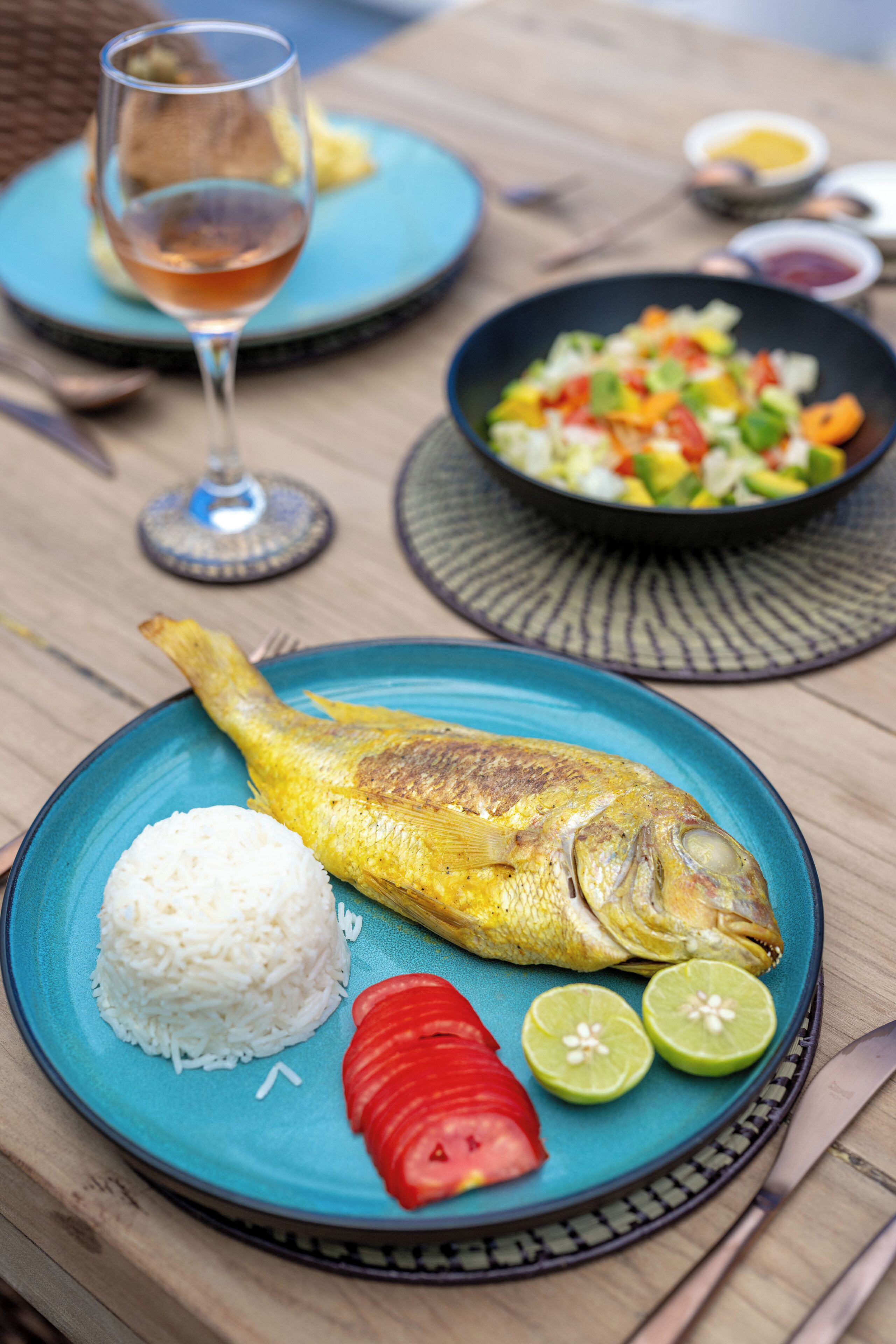 Een bord met vis, rijst en gesneden groenten, een glas drinken en een kom salade op een tafel.