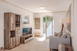 Un salon avec un canapé gris, un meuble en bois, une télévision et une porte donnant sur l’extérieur.