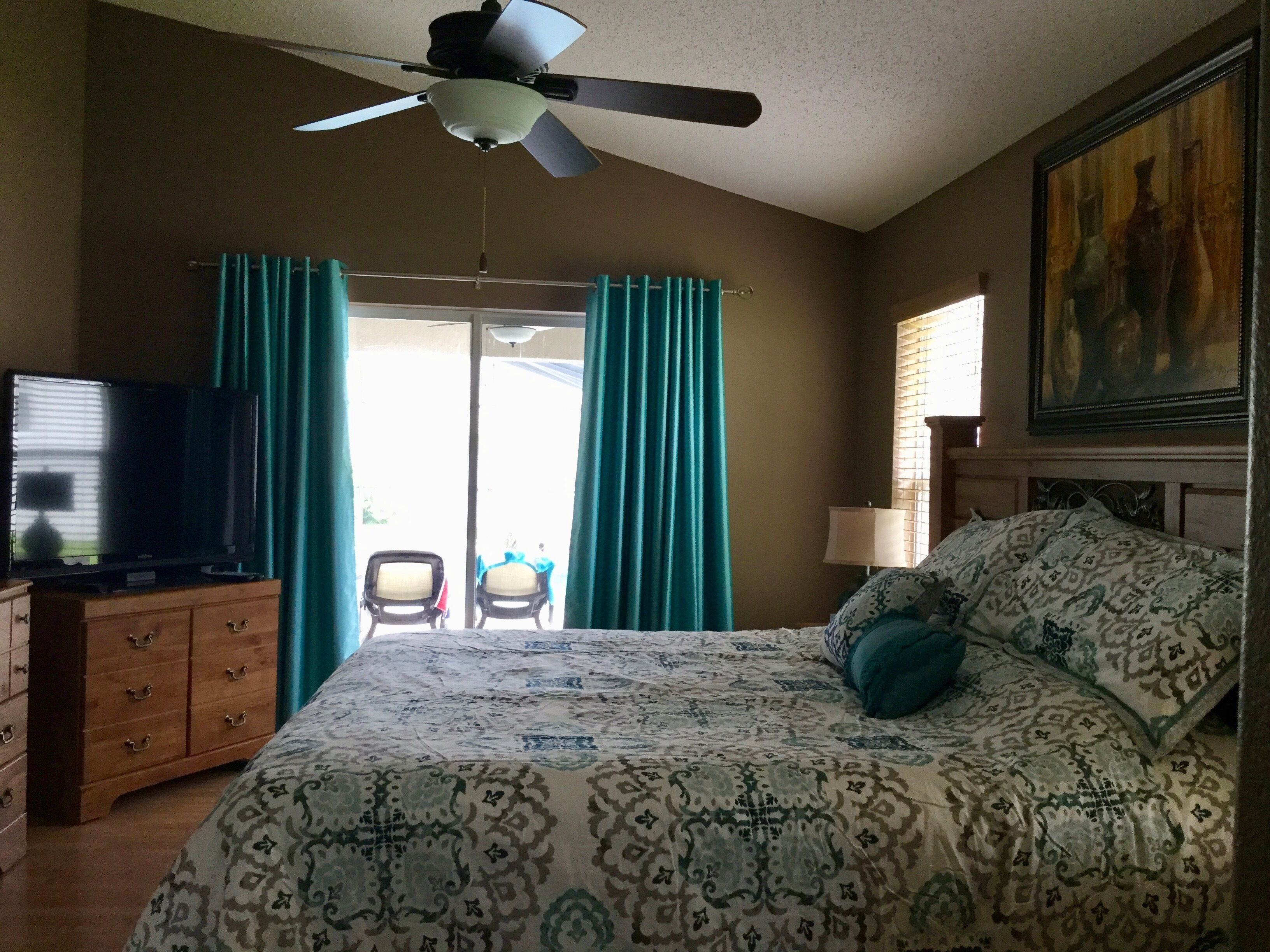 A bedroom with a bed, a ceiling fan, a television, a dresser, and a painting on the wall.
