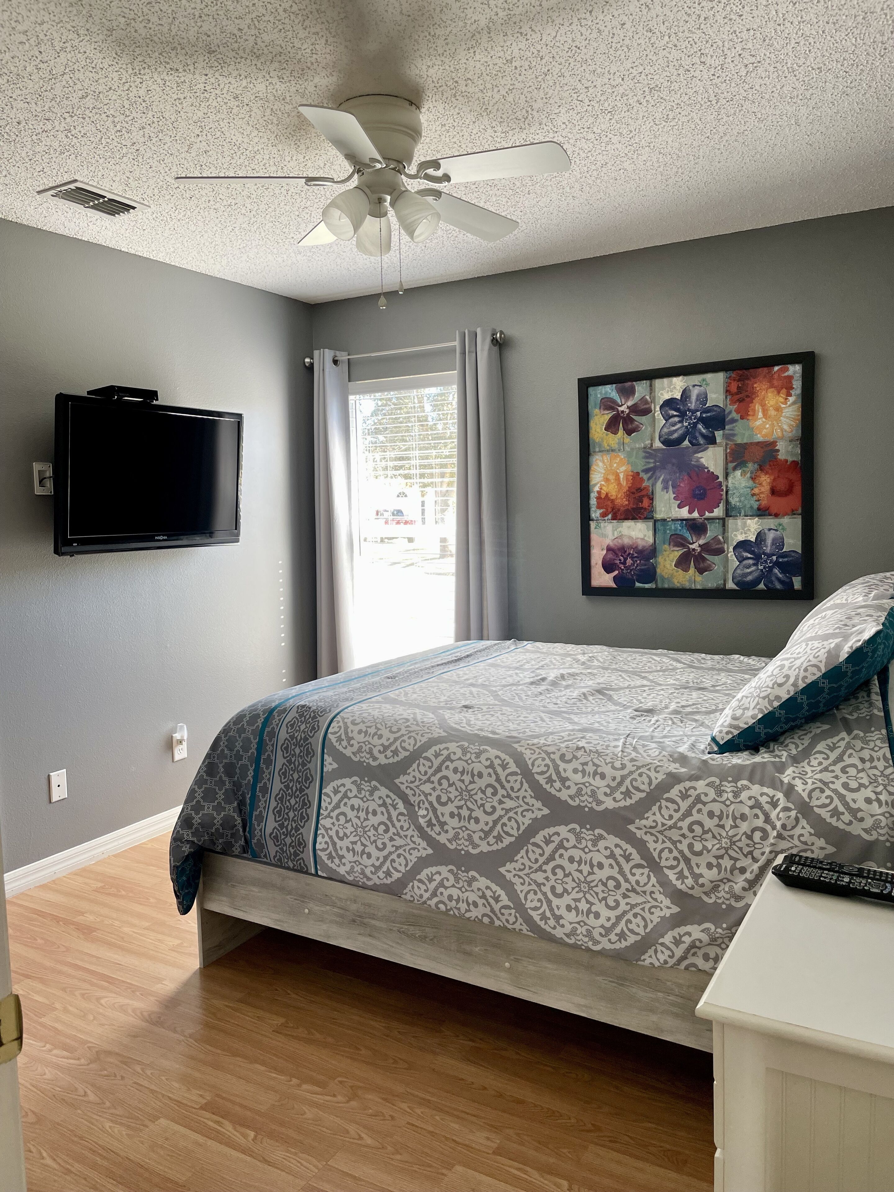 A bedroom with a bed, a ceiling fan, a TV mounted on the wall, and a window with curtains.