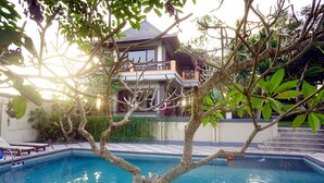A swimming pool with a tree in the foreground and a two-story building in the background. - Villa Annapurna Bali (Mengwi)