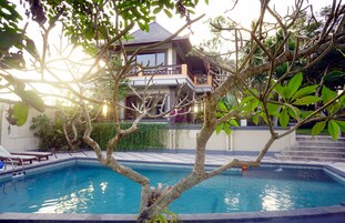 Una piscina con un árbol en primer plano y un edificio de dos plantas al fondo.