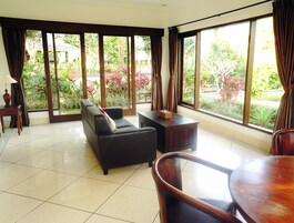 Amplia sala de estar con sofá, mesa de centro y mesa de comedor. Grandes ventanales ofrecen vistas a un jardín con diversas plantas y árboles.