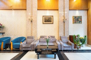A lobby with a reception desk, two blue armchairs, a grey sofa, a black coffee table, and a vase with flowers. - Lucky Star Hotel 146 Nguyen Trai (Ho Chi Minh City)