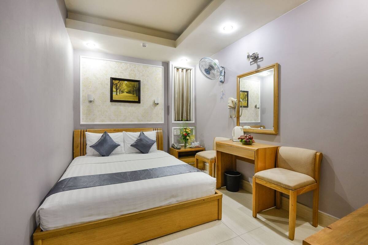 A bedroom with a bed, bedside table, chair, mirror, and a wall-mounted fan.