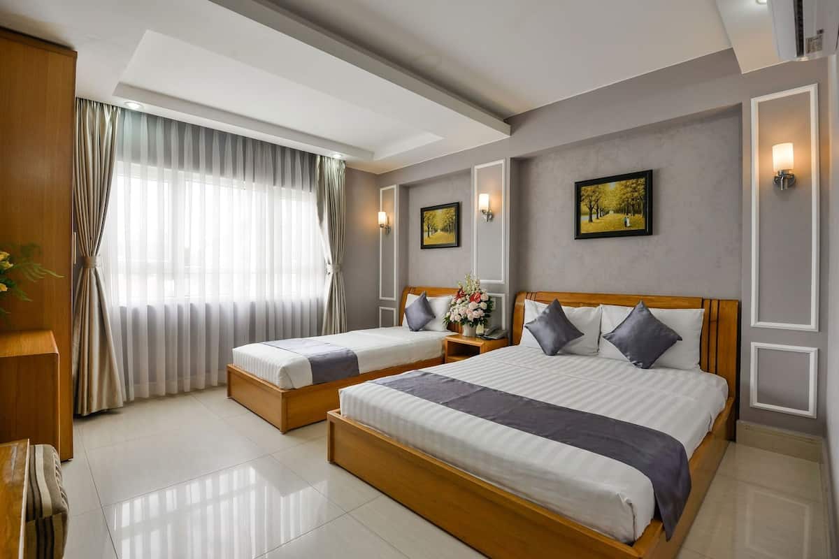 A hotel room with two beds, a wooden headboard, a large window with sheer curtains, a nightstand with a lamp, and framed artwork on the walls.