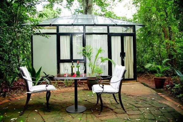A conservatory with a glass roof, two white chairs, a table with wine bottles, and potted plants. - Entim Sidai Wellness Sanctuary (Nairobi)
