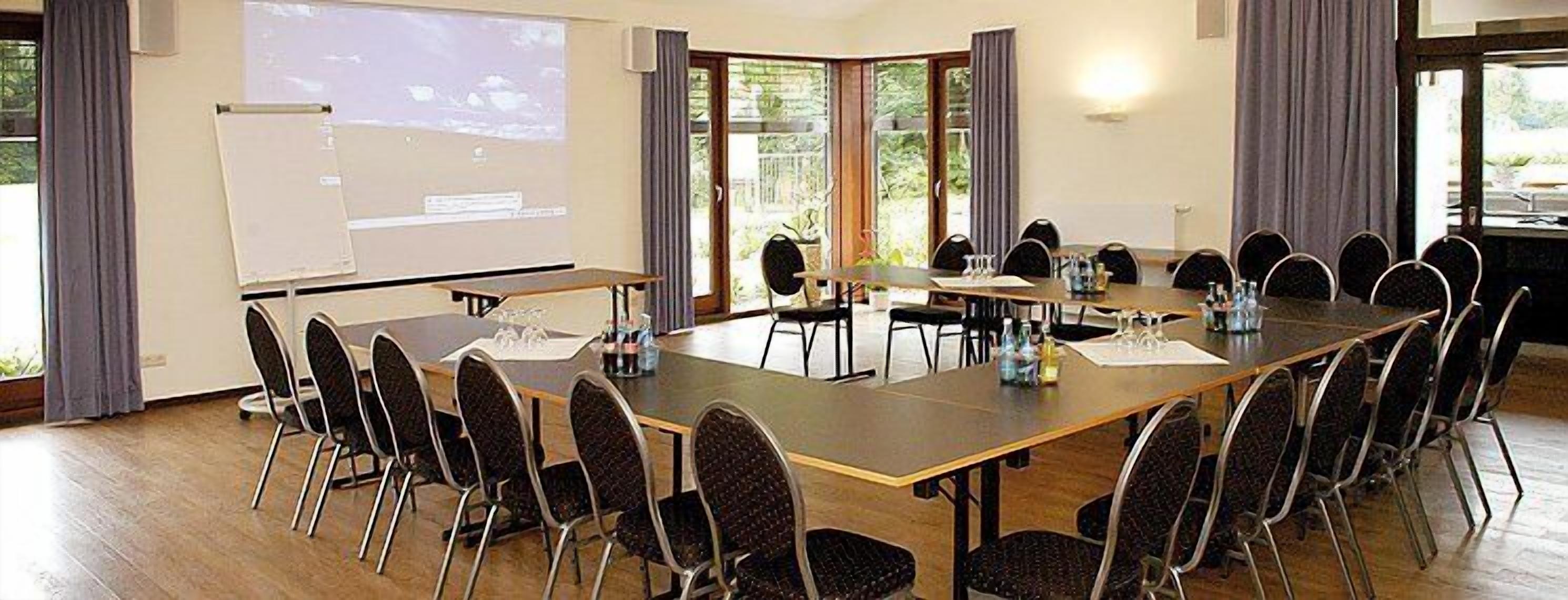 A conference room with a long table, multiple chairs, a projector screen, and large windows with curtains.
