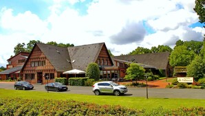 A traditional building with a steeply pitched roof, a central entrance, and a well-maintained lawn with parked cars. - Hotel Landhaus Pollmeyer (Friesoythe)