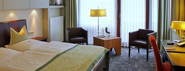 Quarto de hotel com uma cama grande, duas cadeiras, uma mesinha e um abajur.