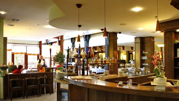 A bar area with wooden furniture, a counter with a bottle of champagne, and a view of the dining area. - Hotel Landhaus Pollmeyer (Friesoythe)