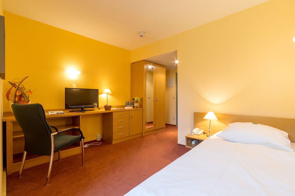 A hotel room with a bed, a desk with a chair, a television, and a nightstand with a lamp.