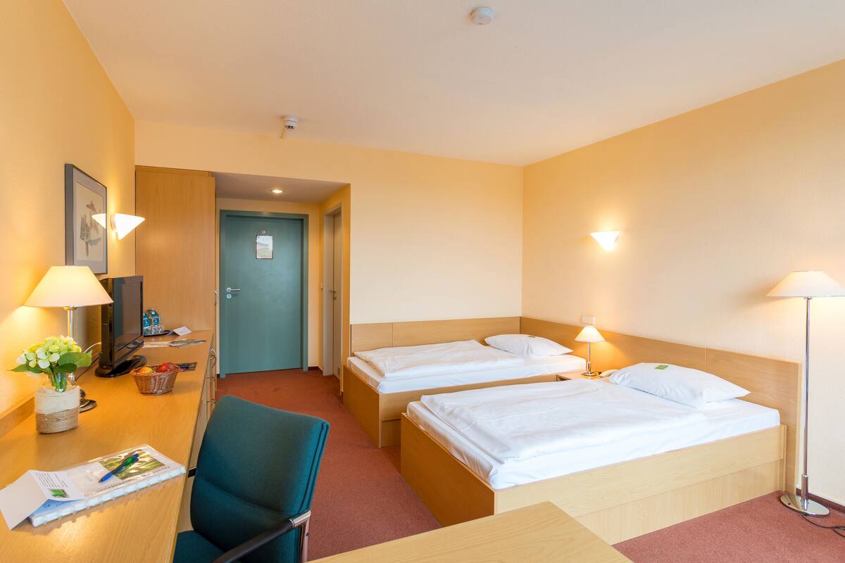A hotel room with two single beds, a desk with a chair, a lamp, a basket with fruits, and a door to the bathroom.
