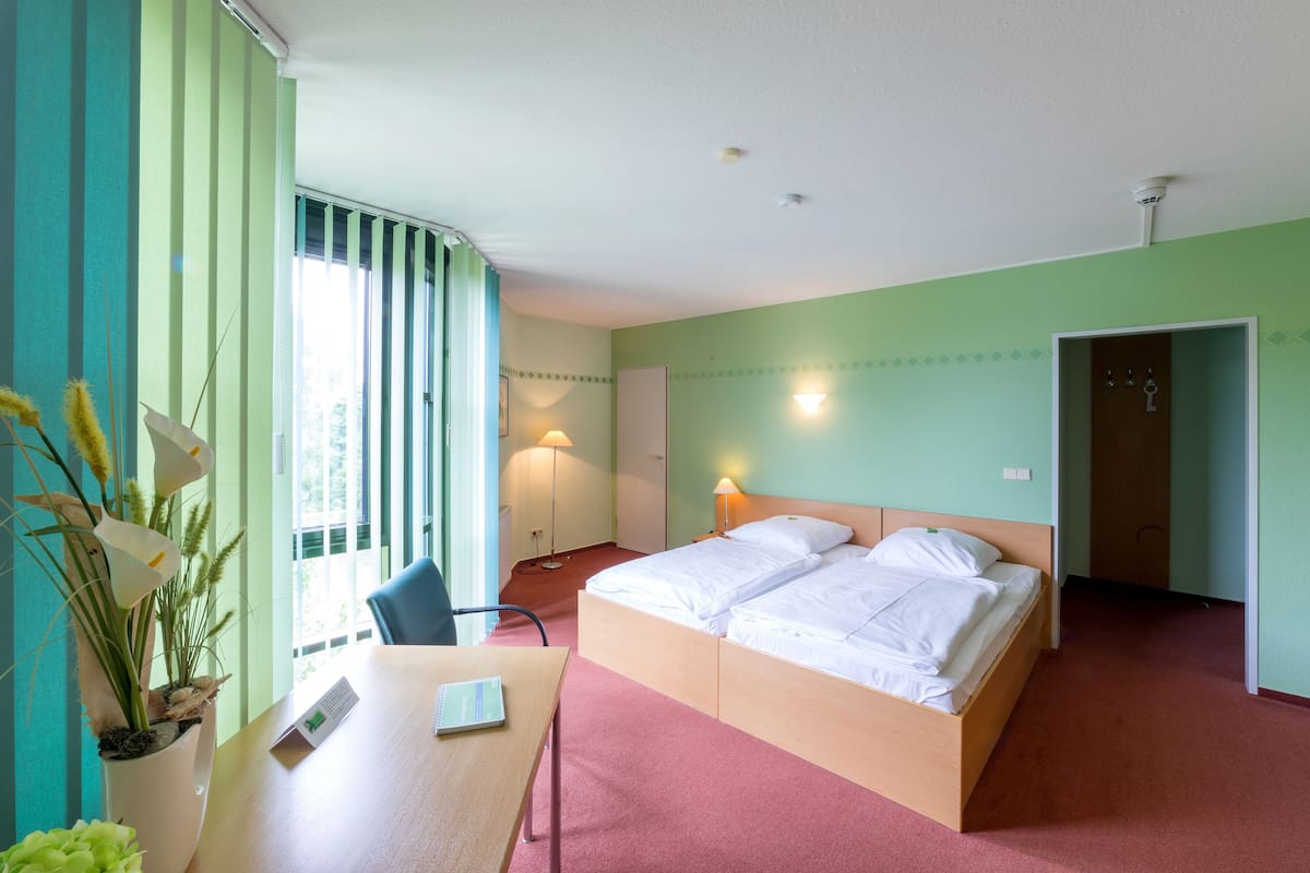 A hotel room with a bed, a desk, a chair, a potted plant, and a window with curtains.